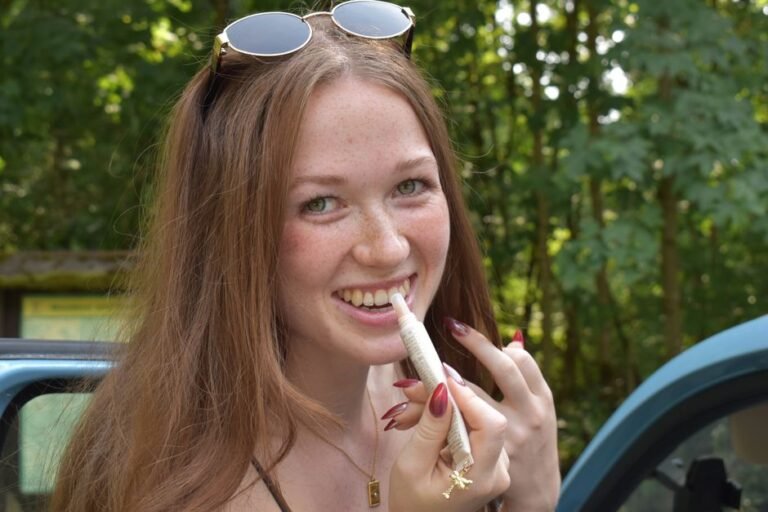 A person in a winter coat standing outside on a sunny day, applying lip balm from a small tube