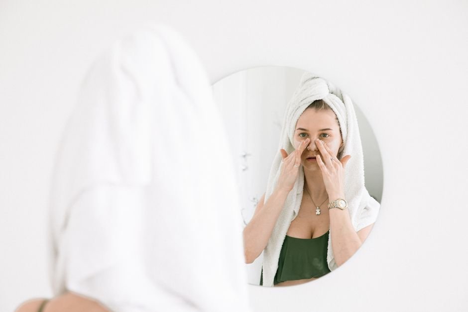 A person standing at a bathroom mirror in the morning light, smoothing sunscreen onto their cheek