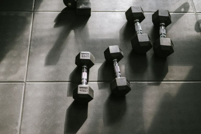 A person kneeling on a gym floor between a set of dumbbells and open floor space