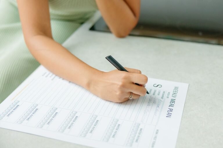 A person sitting at a kitchen table writing in a weekly paper planner next to a mug