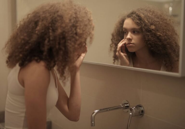 A person standing at a bathroom sink, leaning toward the mirror to look closely at their cheek
