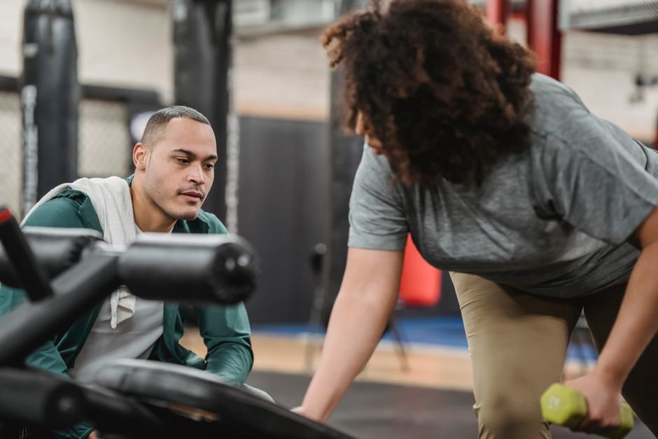 A beginner using gym equipment while a trainer offers guidance nearby