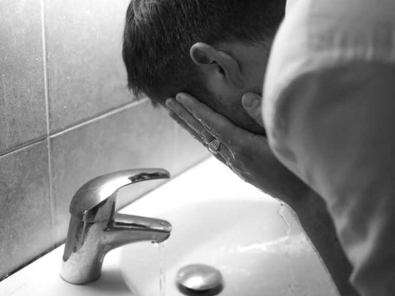 A person rinsing their face with water over a bathroom sink, a cleanser bottle on the counter beside them