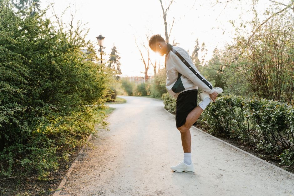 A runner in ordinary workout clothes stretching their calf against a low wall beside a park path